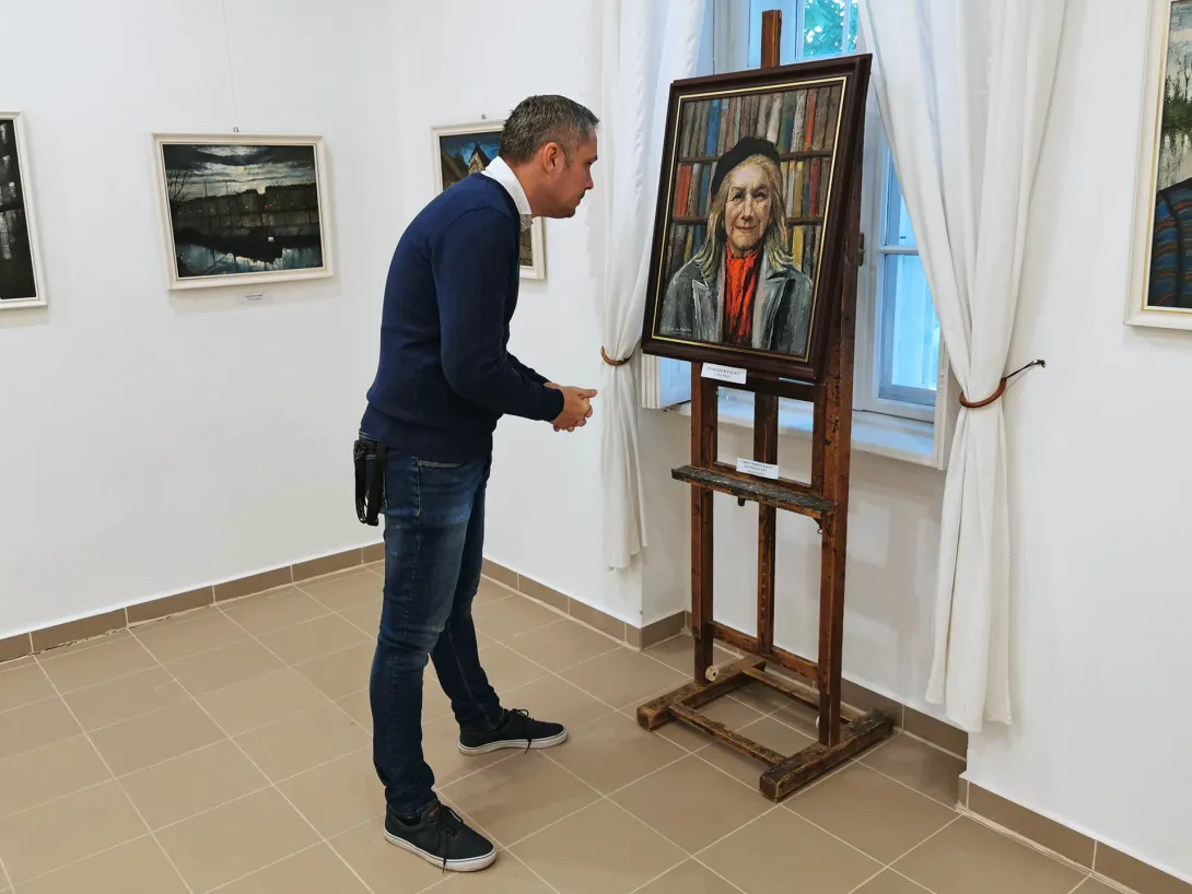 Önarckép 80 évesen. Idős nő portréja könyvespolc előtt. Fekete sapkát, piros sálat és szürke kabátot visel., Corini Margit festőművész születésének 125., halálának 40. évfordulójára rendezett emlékkiállítás, Vidovszky Béla Városi Képtár, Gyomaendrőd