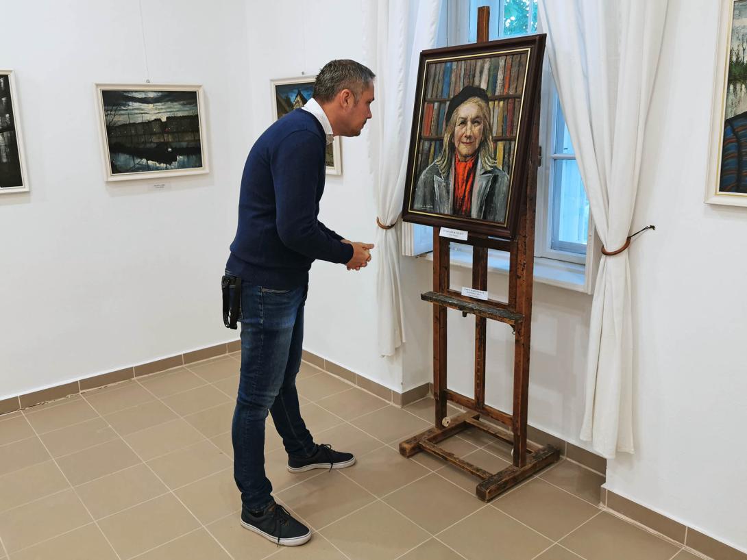 Önarckép 80 évesen. Idős nő portréja könyvespolc előtt. Fekete sapkát, piros sálat és szürke kabátot visel., Corini Margit festőművész születésének 125., halálának 40. évfordulójára rendezett emlékkiállítás, Vidovszky Béla Városi Képtár, Gyomaendrőd